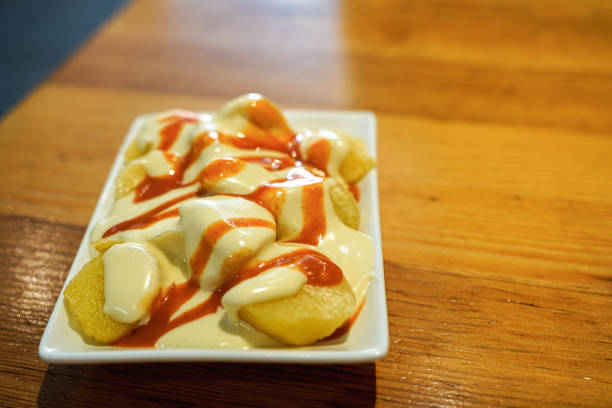 On the wooden table a plate with a portion of patatas bravas, typical Spanish ration, potatoes in two sauces, mayonnaise and spicy red sauce, no one is seen.