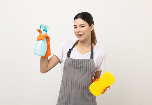 Young beautiful woman with apron and rubber glove ready for cleaning home on isolated white background. Housekeeping housework or maid worker holding spray bottle and sponge