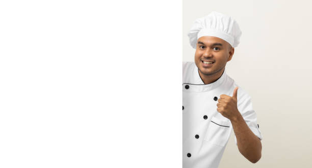 istock Happy Young handsome asian man chef in uniform looks out from behind an empty whiteboard. Cooking indian man hiding behind big blank billboard for advertising text menu in kitchen and restaurant. 1406693824