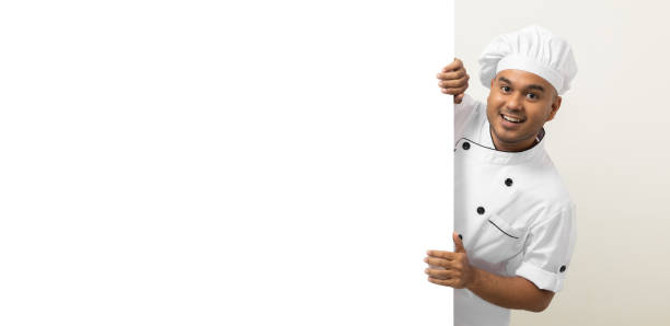 istock Happy Young handsome asian man chef in uniform looks out from behind an empty whiteboard. Cooking indian man hiding behind big blank billboard for advertising text menu in kitchen and restaurant. 1406694185