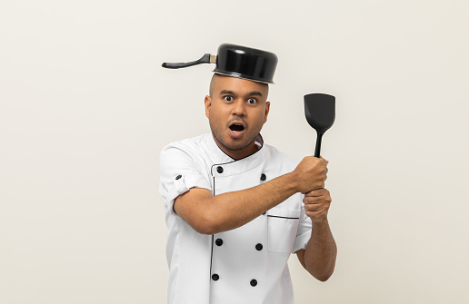 Funny excited face Young asian man chef in uniform put a stew pot over the head holding turner kitchen utensils on isolated. Cooking indian man Occupation chef People in kitchen restaurant and hotel.