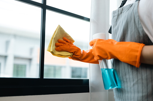 Beautiful housewife with rubber gloves and apron using spray bottle and microfiber towel to clean windows at apartment. Young woman is happy to clean home. Maid cleaning service.