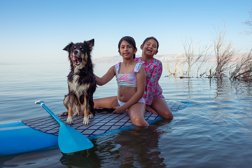 Two happy girls and their dog having fun with a stand-up paddleboard (SUP) on vacation on the lake in summer. They are posing for the camera, sitting on a surfboard, and floating on the water. The dog is mixed-breed Husky and an Australian shepherd obediently stays near them. One of the girls is petting the dog.