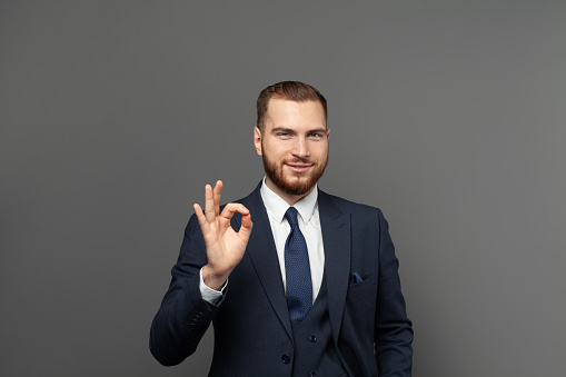 Happy young businessman showing ok sign over grey background