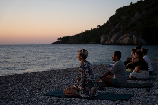 Group of people meditating on the beach at sunrise, practicing yoga for mental health