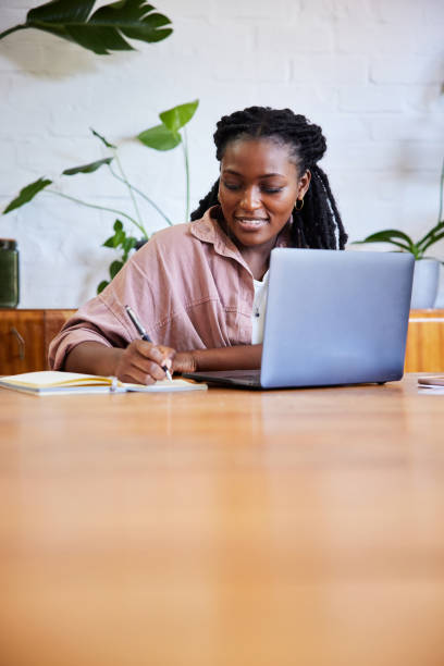 istock Smiling young woman writing notes and using a laptop while working from home 1406698277