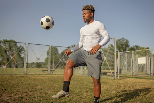 A young African American man is practising his kicks with a soccer ball, while standing on a grass field in sports clothing.
