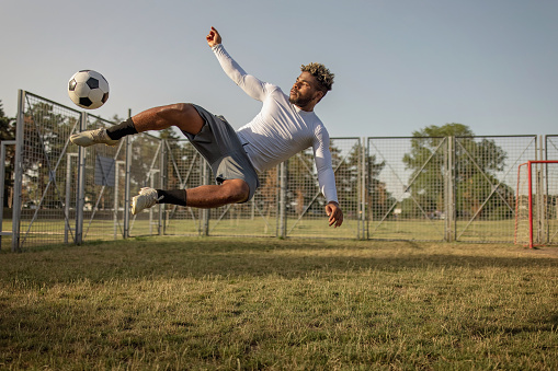 A young African American man wearing sports clothing is jumping in the air while kicking a soccer ball on a grass field.
