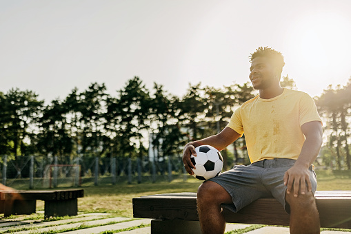 A young African American man is cheerfully sitting on a bench and holding a soccer ball, with the sun behind him.