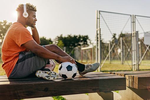 A young African American man is listening to music on his headphones, while resting on a bench with a soccer ball next to him.