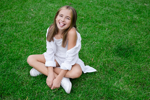pretty tween girl in white clothes sitting on green grass outside in park on sunny summer day. charming ukrainian child portrait.