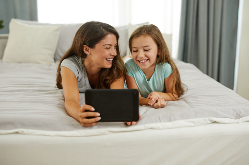 An adorable little girl and her mother using a digital tablet together on the bed at home