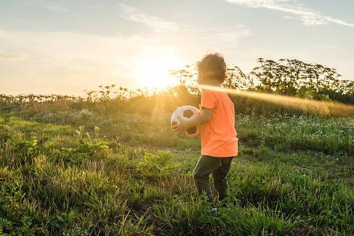 Cute curly Boy with his ball in the field. Beautiful sunset light on background.