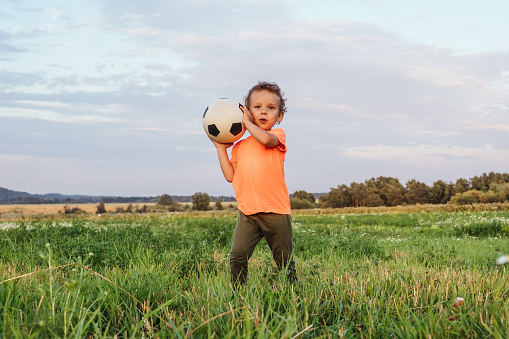 Cute curly Boy with his ball in the field. Beautiful sunset light on background.