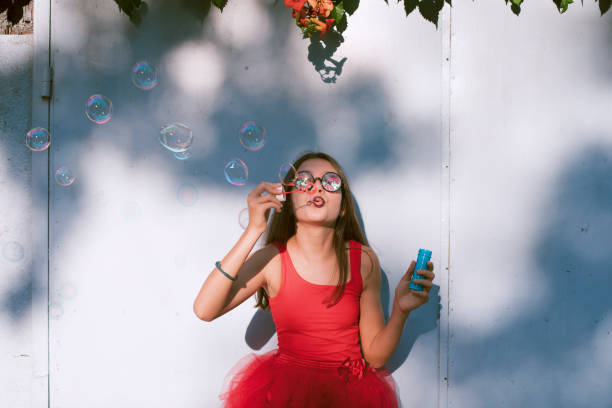 Happy girl having fun blowing bubbles. Dressed in red. Grey background. Summer joy.