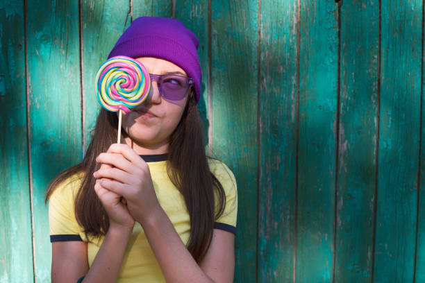 Young woman with long dark hair lying on a yellow blanket, holding a yellow lollipop in her mouth and taking a selfie