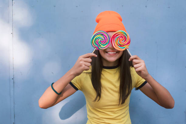 Portrait of a happy girl with lollipops and orange knit hat in front of gray wall.