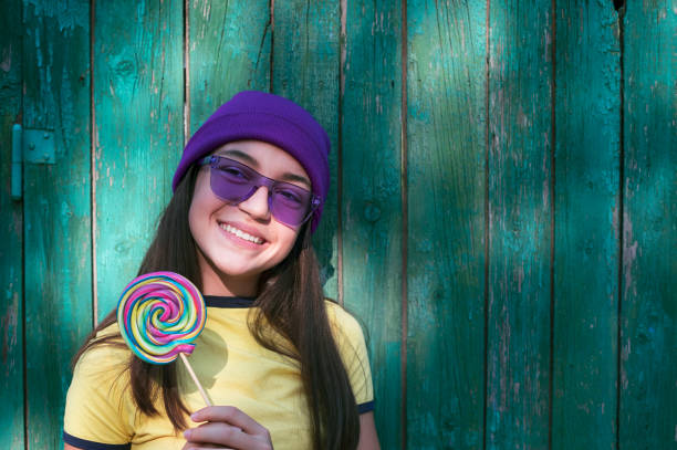 Portrait of beautiful girl with lollipop, purple knit hat and purple sunglasses in front of wooden green background.