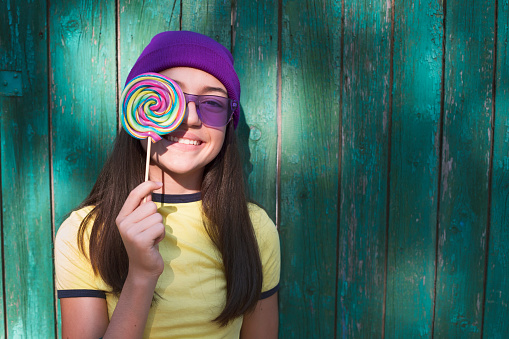 Portrait of beautiful girl with lollipop, purple knit hat and purple sunglasses in front of wooden green background.