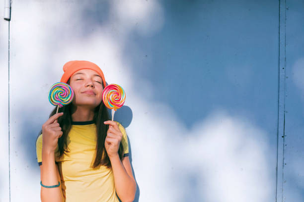 Portrait of a happy girl with lollipops and orange knit hat in front of gray wall.