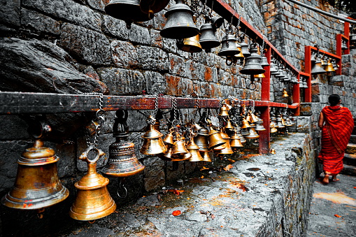 Dakshinkali Temple, Kathmandu, Nepal - April 27, 2022: Nepali woman in red,  leaving the temple, passing by a row of small hanging bells. Most Hindu temples have bells, placed outside, inside, or around the temple. People ring the bell upon entering and leaving the temple. Bells are designed to make a long, sharp sound and there are both scientific and religious beliefs for the effect of that sound.