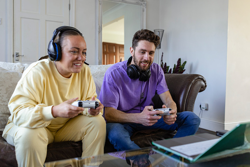 A front-view shot of two friends sitting on the sofa playing a video game together, they are holding video game controllers and wearing headsets.