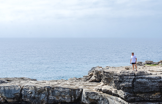old man looking towards the horizon from a cliff on the island of menorca. tourist explores a rocky path on a cloudy day. image conveys calm, happiness. concept of travel destination