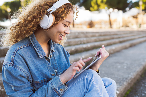 Hispanic woman with blonde curly hair, listens to music with headphones, and uses a tablet pc, in the city