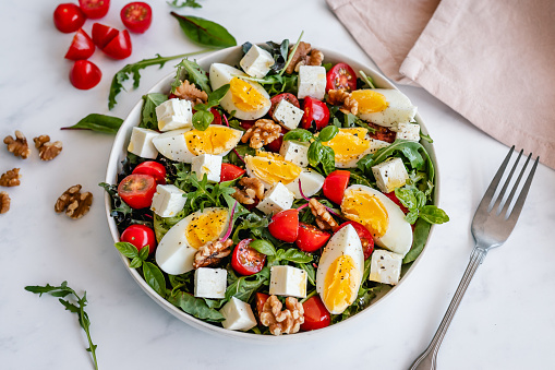 Angled view of a bowl of salad with hard-boiled egg, feta cheese, walnuts and cherry tomatoes, drizzled with olive oil