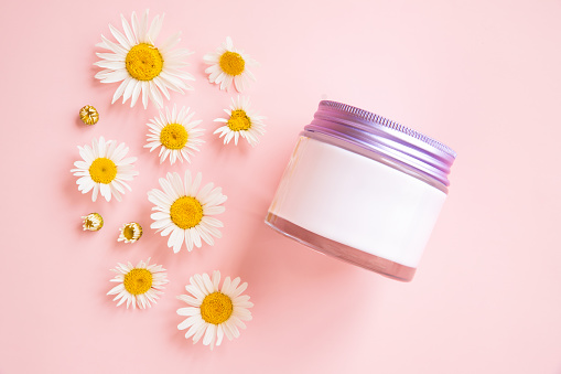 White moisturizing cream in glass jar with metal screw cap and small daisy chamomile flowers on pink background. Mockup, front top view, closeup. Product branding, whitening cream lotion, moisturizer