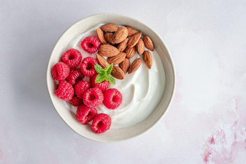 Overhead view of a bowl of yoghurt with raspberries and almonds on a light pinkish background