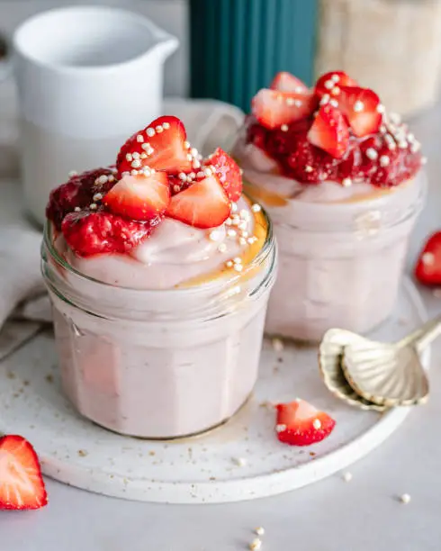 Strawberry mousse topped with strawberry jam and fresh strawberries, drizzled with syrup, in two dessert jars standing on a ceramic platter