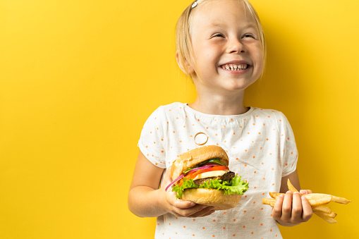 Cute little caucasian girl with blonde hair enjoying burger on a yellow background. Happy kid smiling and eating fast food burger