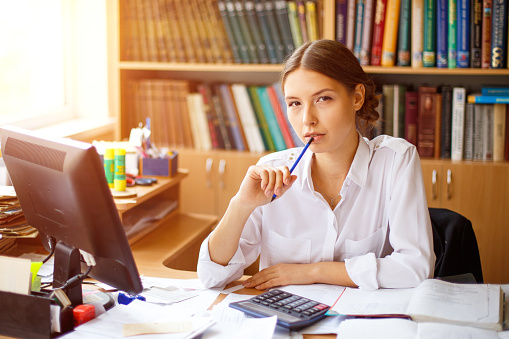 Young woman planning budget, using calculator and computer, reading documents, checking finances, counting bills or taxes, online banking services, sitting at table in office