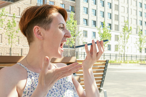 Woman with short hair holds a smartphone near her mouth and screams furiously into the microphone. A woman records a voice message. Quarrel on a mobile phone.