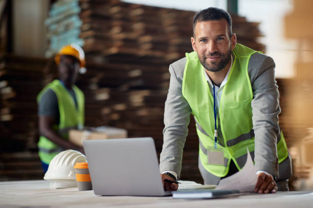 Happy businessman working on laptop at wood warehouse and looking at camera. Happy warehouse inspector working on a computer at looking at camera. foreman stock pictures, royalty-free photos & images