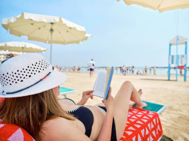 istock Woman wearing a summer hat and lying on a deckchair at the beach while relaxing reading a book, in the background umbrellas, a lifeguard tower and bathers cooling off by the Adriatic sea, Italy. 1406766579
