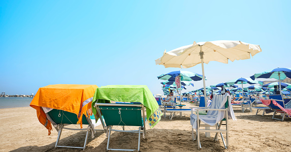 Beach bed with colorful towel in the foreground on a sunny summer day. In the background, the beach equipped with deck chairs and umbrellas on the shores of the Adriatic Sea in Italy.