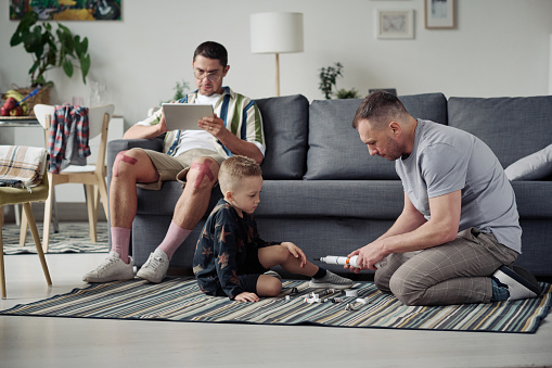Gay dad playing toys with his adopted son on the floor in living room with other dad using tablet pc sitting on sofa in background