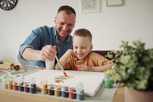 Happy kid boy learning to paint with paintbrush together with his dad at table, they painting a picture and laughing