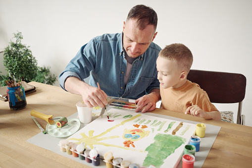 Young father using paintbrush and paints to help his little son to paint a picture on paper at table