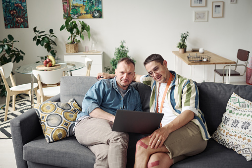Homosexual couple watching movie online on laptop while sitting on sofa in living room