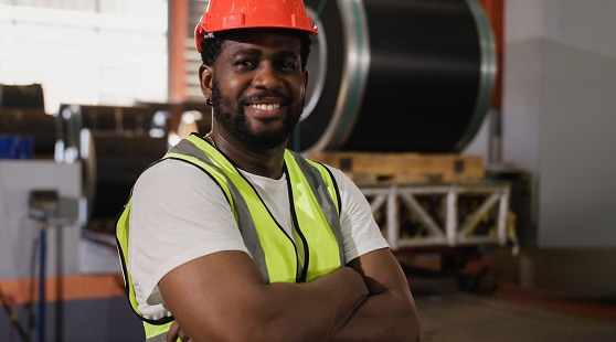 Portrait of happy professional African American male engineer wearing safety helmet standing and arms crossed in factory industry. Male worker confident smiling and looking at camera in a warehouse.