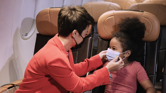 Young mother wearing protective face mask to her daughter for protection coronavirus disease or covid-19 in cabin of airplane. Little African child girl with afro hair and her mom wear mask in flight.