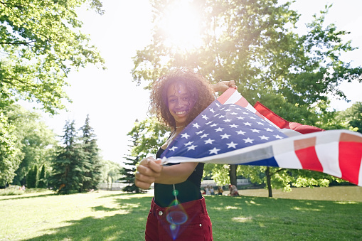 Young African american woman is waving American flag on top of at nature background. USA resident, US citizen. Immigration concept