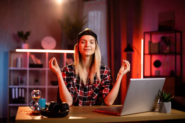 piacevole donna che medita con gli occhi chiusi mentre è seduta alla scrivania con un laptop moderno. - yoga business women living room foto e immagini stock