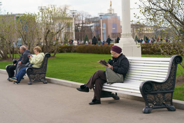 Moscow, Russia - May 3, 2022: Senior woman sitting on the bench in the public park VDNkH. She is reading newspaper. Other senior woman using smartphone, looking at the screens. Lifestyle concept.