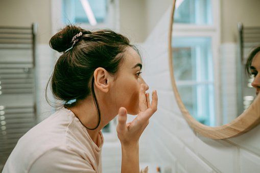 Photo of a young woman getting ready in the bathroom for the upcoming day; applying moisturizer as a part of her daily routine.