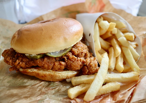 A fast food fried chicken sandwich is pictured (with selective focus) nestled next to a side of fries.