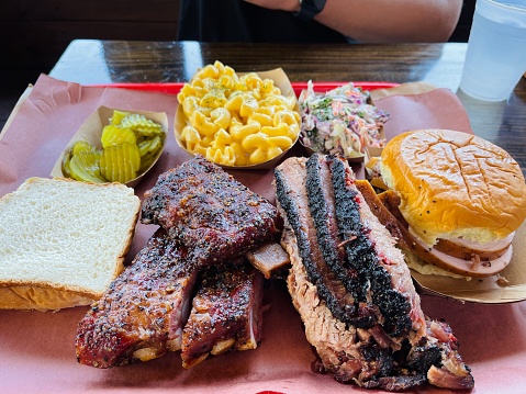 A large platter of barbecue meats and sides, including pork ribs, beef brisket, smoked turkey, mac and cheese, cole slaw, pickles, and bread, from a local restaurant in Austin, Texas, USA.